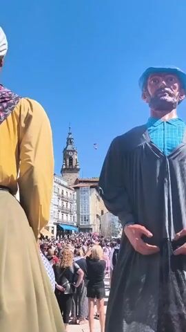 Two giant figures, one in a yellow top and green skirt, the other in a grey robe and blue beret, stand in the foreground. In the background, a crowd of people gathers in a square. A clock tower with a pointed spire rises above buildings, and a statue of an angel is visible. A small object, possibly a person in a parachute or a small carriage, is suspended in the air by a wire between two points. The sky is clear and blue.
