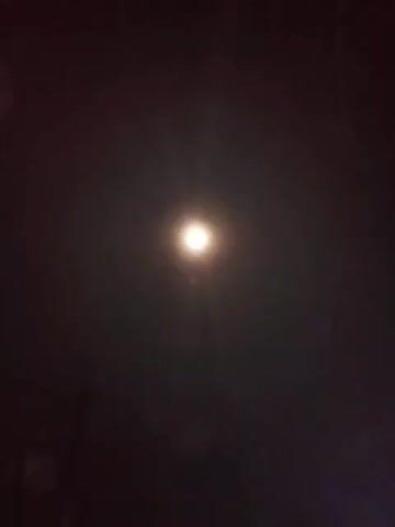 The video shows a night sky with a bright full moon, which is surrounded by some hazy clouds. The camera moves slightly, shifting the moon's position in the frame. There are also glimpses of dark shapes that could be trees or buildings in the bottom part of the frame. The overall environment is dark, with the moon providing the primary source of light.