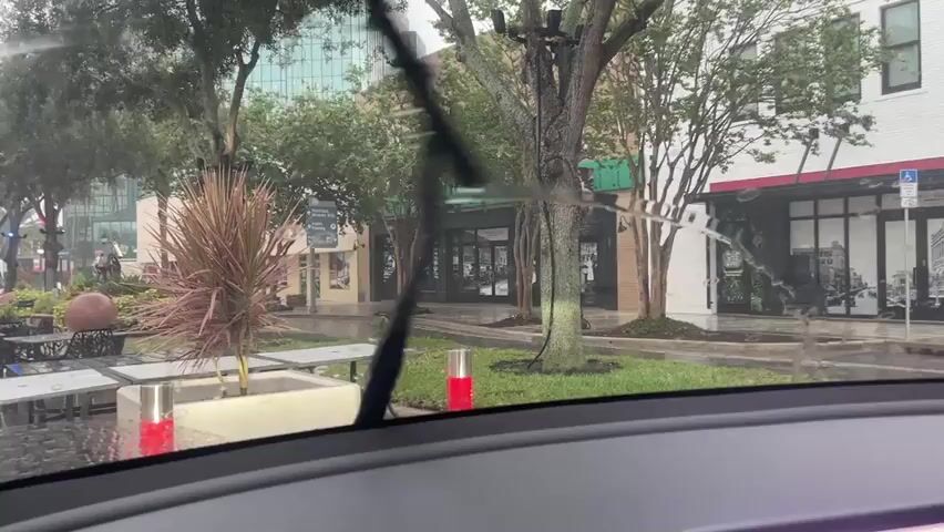 The video shows a view from inside a vehicle looking out through the windshield. It is raining, and the windshield wipers are active, clearing streaks of water from the glass. The scene outside is a street with businesses and trees. There are tables and chairs set up outside one of the businesses, suggesting an outdoor dining or seating area. The buildings appear to be made of brick and have large windows. The sky is overcast, and the lighting suggests it is daytime. There are no people clearly visible in the frame. The overall environment appears to be a commercial or urban area experiencing inclement weather.