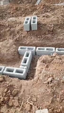 The video shows a construction site in a rural, possibly arid, area. The ground is dry and mostly brown with scattered loose dirt and small rocks. Numerous rectangular grey concrete blocks are scattered around the site. Some blocks are stacked in piles, while others are laid out in trenches, forming the foundation of a building. The blocks have large rectangular holes in them. In the distance, there are other structures that appear to be under construction or partially completed. The video seems to show the early stages of building construction.