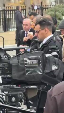The video shows a crowd of people, many of whom appear to be media professionals due to the presence of professional video cameras.  The cameras are mounted on tripods and covered with black protective bags.  Several individuals are visible in the background, some wearing coats and hats, suggesting a cool climate. One person is drinking from a cup. The setting appears to be an outdoor area near a fence or gate. The scene seems to be associated with a news event or media gathering.