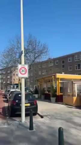 The video shows a street in what appears to be a European city. The camera starts by focusing on a street sign indicating a "Zone 30" speed limit.  A yellow-walled business with outdoor seating is visible in the background.  The camera then pans to the left, revealing a red-brick bike lane alongside a road with parked cars and multi-story apartment buildings on either side.  A few bare trees are scattered along the street. The sky is clear and blue. A pigeon walks across the bare soil between the bike lane and the parked cars.