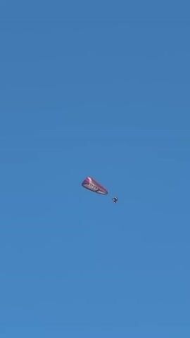 A paraglider with a pink wing flies through a clear blue sky. A person is suspended beneath the wing. The paraglider moves slowly across the frame.
