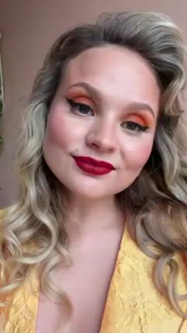 A woman with blonde, curly hair and bright red lipstick is standing in front of a beige wall. She is wearing a yellow brocade dress. Her makeup includes orange eyeshadow and black eyeliner. She holds up a peace sign with her right hand, smiling.