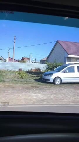 The video shows a view from a vehicle as it drives past various buildings and landscapes. The first scene shows a small, light-colored building with a dark roof, possibly a shop or a small business, with a vending machine in front. A white van is parked to the left. The next scene reveals a rural landscape with several houses, some with distinctive architectural styles, and trees. The final scenes continue to show rural settings with more buildings, including what appears to be a farm or industrial complex with large structures and silos. The sky is clear and blue throughout the video. The footage is slightly blurry, suggesting it was filmed while the vehicle was in motion.