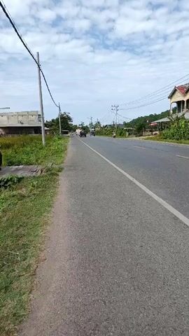 This is a vertical, first-person perspective video recorded on a cloudy day. The video begins by showing a paved road with a grassy shoulder on the left side. A power line runs diagonally across the top of the frame. A utility pole is visible on the left side of the road. In the distance, a few vehicles can be seen on the road, including what appears to be an older model Jeep. Buildings are visible on the right side of the road, and vegetation lines the roadside. As the video progresses, the Jeep moves further down the road and passes by another vehicle. A black car is seen traveling in the opposite direction of the Jeep. The video continues with a view of the road ahead, with more vehicles appearing and disappearing from view.