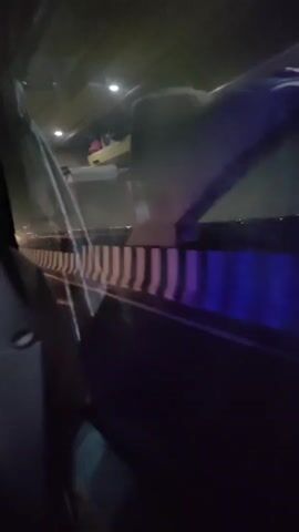 The video shows the interior of a vehicle at night, possibly a bus or train, as seen from a passenger's perspective. The view is partially obstructed by a car window. Outside the window, there are lights indicating a city or populated area in the distance. The vehicle appears to be on an elevated structure, possibly a bridge or overpass, given the railing visible in the foreground. There are reflections on the window, including purple and blue lights, which could be from the vehicle's interior or exterior lighting. In the background, beyond the railing, some distant lights are visible. The lighting conditions are dim, suggesting it is nighttime.