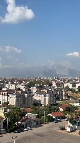 A vertical video shows an aerial view of a city skyline with mountains in the distance. The sky is blue with scattered white clouds. Buildings of various heights and architectural styles fill the mid-ground and foreground. Some buildings are multi-story apartment complexes, while others appear to be smaller houses with red-tiled roofs. Green trees and vegetation are interspersed throughout the urban landscape. Several cars are parked on the street in the foreground. The overall impression is that of a sunny day in a densely populated urban area.