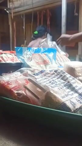 The video shows a close-up of packaged food items in a green tray. In the background, a person wearing a blue shirt is visible, and another person wearing a pink shirt with a green clip in their hair is also present. They appear to be in a market or food stall setting. The focus is on the food products, with clear labels on some of the plastic packaging. The lighting is dim, suggesting it might be evening or an indoor setting.