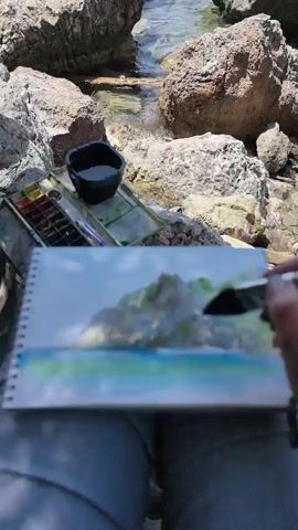 A person is sitting on the rocks by the sea, painting a watercolor. The painting is of the sea and a rocky island. The person is holding a brush and painting on a sketchbook. There is a watercolor palette and a cup of water next to the sketchbook. The sea is blue and the rocks are brown and gray. The sky is blue.