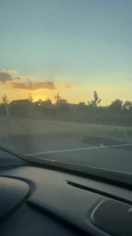 This video captures a sunset view from the interior of a moving vehicle. The camera is positioned to look through the windshield. The sky is a gradient of blue and yellow, with scattered clouds. The sun is visible in the distance, partially obscured by trees and haze. The landscape consists of trees and vegetation along the side of a highway. The road itself is visible, with lane markings. The interior of the car, specifically the dashboard, is also visible in the foreground. A black car passes by in the middle ground. The overall impression is a scenic drive during golden hour.