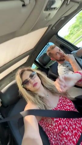 The vertical shot captures two people inside a car, presumably a couple, enjoying a drive during bright daytime in Mahala, Montenegro. The blonde-haired woman in the foreground is taking a selfie video or photo, smiling widely with her mouth open, and gesturing animatedly with her right hand, suggesting she's singing or dancing to music. She wears dark cat-eye sunglasses, a red spaghetti-strap sundress with a white floral pattern, a silver necklace, and a red beaded bracelet on her left wrist. Her black seatbelt is securely fastened across her chest.

The man, presumably the driver, is seated in the background. He has short brown hair and a light beard, and is wearing a light grey short-sleeved shirt with a subtle pattern. He is smiling faintly, with his left hand near his chin, and appears to be looking ahead while driving. A black seatbelt crosses his chest.

The car's interior features dark leather seats and a light-colored headliner, with a sunroof partially visible above. The exterior environment, blurred through the windows, shows a clear sky and lush green roadside vegetation, indicating they are on a scenic road. The overall mood is one of happiness and lighthearted enjoyment. There is no visible text in the image.