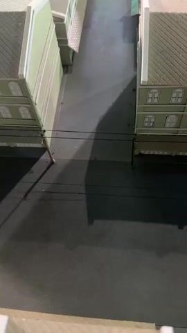 The video shows a miniature diorama of a street scene. The buildings on either side of the street are made of light-colored material, possibly wood or plastic, with green facades on the lower levels and white trim. The roofs are textured to resemble shingles. The street itself is dark gray, suggesting asphalt. There are power lines running across the street, supported by small poles. In the foreground, a shadow is cast across the street, indicating a light source from the left. The camera slowly pans up and away, revealing more buildings and rooftops in the background, with some appearing to be older or more weathered than those in the foreground. The diorama depicts a dense urban or town environment.