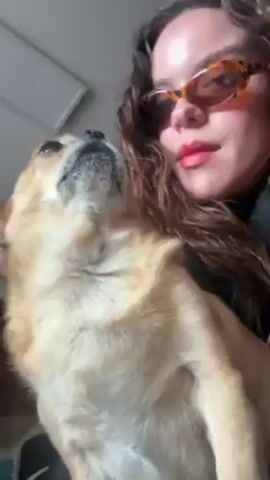 A fair-skinned woman with long curly brown hair and wearing orange-tinted sunglasses and red lipstick holds a light brown chihuahua. The woman kisses the dog's head twice. The dog is looking up at the woman. The setting appears to be indoors, possibly a home. The camera moves slightly, following the actions of the woman and the dog.