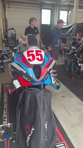 Two young men and an older man are standing in what appears to be a garage or pit area. In the foreground, a highly decorated racing motorcycle with the number 55 prominently displayed on its front fairing is parked on a black mat. A black cover bag is placed in front of the motorcycle. To the right, another motorcycle is visible, with a person kneeling beside it, seemingly working on it. The environment is industrial, with metal shelving and equipment in the background. The lighting is artificial, suggesting an indoor setting. The focus is on the racing motorcycles and the individuals associated with them.