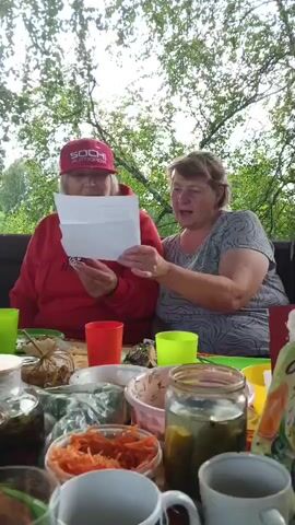 Two women are sitting at a table outdoors, surrounded by trees. The woman on the left wears a red hoodie and a red baseball cap with "SOCHI AUTODROM" written on it in white text. She is holding a piece of paper and reading from it. The woman on the right wears a grey t-shirt with a patterned design and is looking at the paper.

The table is set with various dishes, including jars of pickles and shredded carrots, bowls of food, and plastic cups in green and red. The setting appears to be a backyard or garden, with dappled sunlight filtering through the leaves of the trees. The time of day seems to be daytime, likely afternoon, given the lighting. The overall atmosphere appears relaxed and informal, suggesting a casual gathering or meal. There is no indication of specific weather conditions beyond the presence of trees and foliage. The women's expressions suggest engagement and perhaps enjoyment of whatever is being read.