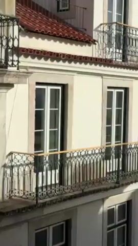 The video shows a light-colored building with white-framed windows and ornate black iron balconies. The building has a red tile roof and is located in a city with other buildings nearby. The sky is blue with some clouds. There are no people visible in the video.