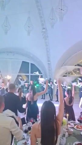 The video depicts a lively indoor event, possibly a wedding reception or a formal party, held in a spacious venue with a high, arched ceiling. The lighting is dim, with warm tones and occasional spotlights creating a festive atmosphere. Several people are visible, mostly in formal attire, dancing and interacting with each other. In the foreground, a woman with long dark hair is seen from behind, wearing a strapless dress, with her right arm raised, holding a white cloth. Another person, a man wearing a white shirt, is seen in the mid-ground, also with his arm raised. In the background, more individuals are dancing, and the venue appears to be set up with tables and chairs, suggesting a dining area. The overall impression is one of celebration and enjoyment.