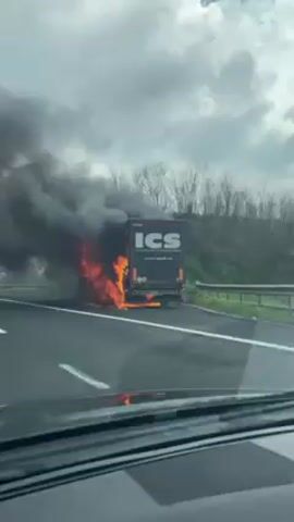 The video shows a car on fire on a road, likely a highway, as seen from another vehicle. Large flames and thick black smoke billow from the burning car. The road has lane markings and appears to be relatively flat. The sky is overcast.