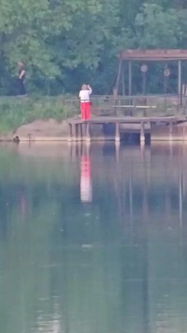 The video shows a tranquil lakeside scene, likely captured on a calm day. In the middle ground, there's a wooden dock extending into the water with a small structure on it. A person wearing a white top and red pants is standing on the dock, facing away from the camera, seemingly looking out over the water. Their reflection is visible in the lake. Lush green trees border the lake on the opposite side. In the distance, you can see hints of houses and cars, indicating a populated area nearby. The camera zooms out, revealing a portion of a gray wooden railing in the foreground, suggesting the video was taken from another dock or platform. The overall impression is serene, with the still water providing a mirrored reflection of the landscape.