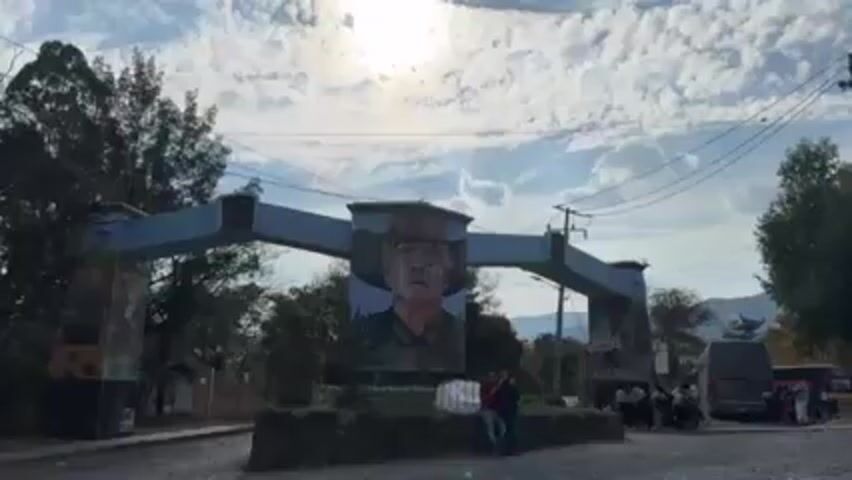 The video shows an archway painted pale blue with a large portrait of a man wearing a hat. The portrait is centered on the archway, and the sky is visible behind it, with some trees and mountains in the background. There is a group of people standing near a bus to the right of the archway. Two people are standing to the left of the archway. The video zooms in on the portrait.