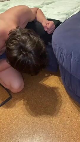 A person is kneeling on the floor, with their head down, appearing to be looking or reaching under a blue couch. The person is wearing black pants and no shirt. The floor appears to be cork or linoleum. There is a shadow on the floor cast by the person and the couch.