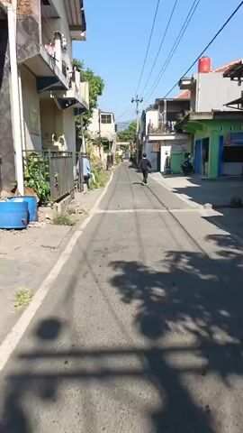 A person is walking down a narrow, paved street lined with houses. The street is in good condition, with white lines marking the edges. There are trees and vegetation on either side of the street, and power lines run overhead. The sky is blue and clear, suggesting it is a sunny day. The person is wearing dark clothing and a backpack, and is walking away from the camera. The houses are mostly two-story buildings, some with balconies. There are motorcycles parked in front of some of the houses. The overall impression is of a quiet residential street in a warm climate.