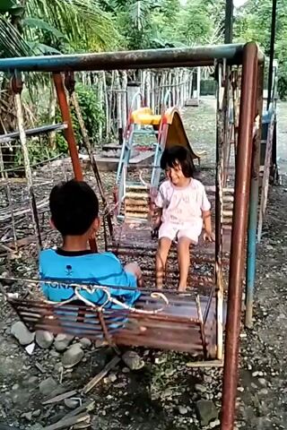 Two children, a boy and a girl, are sitting on a swing in what appears to be a backyard or park setting. The boy, wearing a blue long-sleeved shirt with the name "RONALDO" and the number 7 on the back, is facing away from the camera. The girl, dressed in a light pink outfit, is facing forward. Both are sitting on a weathered, multi-person swing with a wooden slat seat, suspended from a rusty metal frame.

Behind the swing set is a playground with a small slide and ladder. The ground is covered in gravel and rocks. Green foliage and trees surround the playground, suggesting a natural environment. The children appear to be engaged in a calm activity on the swing set.