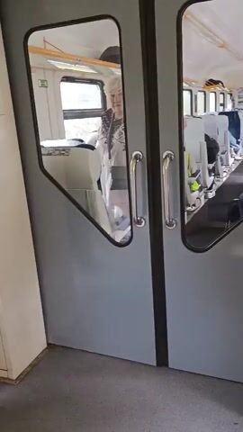 The video shows the interior of a train car. The camera is looking through a set of doors that open inwards. In the foreground, the doors are closed, with handles visible. Through the glass panels of the doors, the interior of the train car can be seen, with rows of seats, some occupied by passengers. The aisle of the train car is visible, and there is a woman standing near the doors. The video then transitions to a closer view of the woman standing near the window of the train door. She is wearing a denim jacket and appears to be looking out the window. The text "НЕ ПРИСЛОНЯТЬСЯ" is visible on the window.