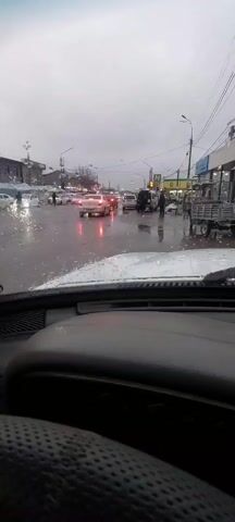 The video is taken from inside a car on a rainy day. The windshield has raindrops on it, and the wipers are turned on. The street is wet and reflects the headlights of other cars. There are several other vehicles on the road, including cars, vans, and a small truck. People can be seen walking along the sidewalks and crossing the street. Buildings and streetlights are visible in the background. The sky is overcast.