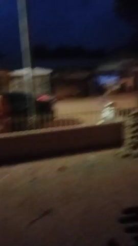 The video shows a street scene at dusk or night. A fence runs across the foreground, separating the viewer from a dirt road. Beyond the road are several buildings, possibly shops, with some lights on. There are a few people and vehicles visible, including what appears to be a scooter. The overall impression is a quiet, somewhat dim, possibly rural or semi-urban environment.