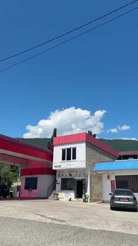 The video opens with a shot of a red and white gas station under a clear blue sky with white clouds. The gas station features a red canopy and white support pillars. To the right of the pumps, a small, two-story building, also red and white, has visible text including "МАГАЗИН" (store), "КОРМА ДЛЯ ЖИ" (feed for animals), and a "Purina" sign. Lush green mountains are visible in the background. The camera then pans left, revealing a dense green landscape with various trees and bushes. Several utility poles and power lines cross the frame. Tall, slender evergreen trees (likely cypresses) are prominent among the foliage. The scene depicts a bright, sunny day in a mountainous, vegetated area.