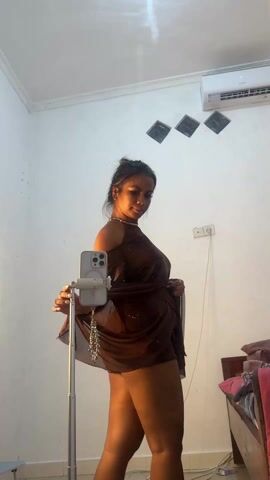 A woman stands in a room in front of a mirror attached to a phone tripod. She has her hair in a bun and is wearing a brown outfit consisting of a short skirt, a halter top, and flowing sleeves connected to the top. A chain is visible across her back. The room has white walls, with an air conditioner and three diamond-shaped decorations. A bed is visible in the background to the right. The woman rotates 180 degrees, showing off her outfit from the back.