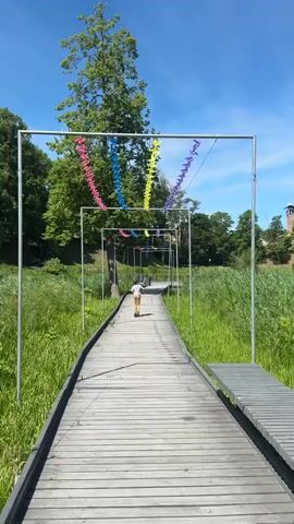 A wooden boardwalk extends forward, flanked by tall green grass. A young child wearing a white shirt and brown shorts rides a scooter down the boardwalk away from the camera. Metal archways are spaced along the boardwalk, adorned with strings of colorful pinwheels—pink, blue, yellow, and purple. These archways create a tunnel-like effect, drawing the eye towards the horizon. Beyond the boardwalk, trees with lush green foliage and a clear blue sky with sparse white clouds form the background. To the right, a section of a brick building is visible amongst the trees.