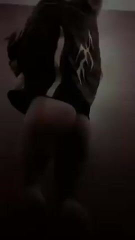 A person in a black jacket, light-colored tights or leggings, and a pink hoodie is dancing or moving erratically in a room. The room appears dark and has a patterned wall. A light fixture is visible on the ceiling. The person's face is obscured by the hoodie in most of the video.
