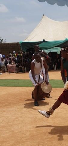 This media file captures a vibrant cultural performance in Unguwar Saya, Nigeria. The scene is set outdoors, under a bright sky, suggesting it's daytime. A large white tent provides shade for spectators in the background, while the foreground is a sandy, brown surface.

The primary subjects are a group of people participating in a ritualistic dance. At the center, a woman in a green top and a maroon skirt is actively engaged, holding a whisk and a drumstick. She is adorned with a beaded headdress and what appear to be decorative anklets. Two men, dressed in white sleeveless shirts and white shorts, are also central to the activity. One man is playing a large drum, and the other is holding a smaller drum and a white feathered object. Both men are barefoot. Further back, other individuals are visible, some holding drums and participating in the performance, while a seated audience watches from under the tent.

The activity appears to be a traditional dance or musical performance, possibly part of a celebration or ceremony. The energy of the performers, particularly the central woman, suggests a lively and engaging event. The expressions on the performers' faces are difficult to discern clearly, but their body language conveys involvement and movement.

Notable details include the traditional attire of the performers and the instruments they use, specifically the drums. The decorative elements on the woman's headdress and anklets add to the cultural richness of the scene. The weather appears clear and sunny, contributing to the outdoor setting. There is no visible text in the media file.
