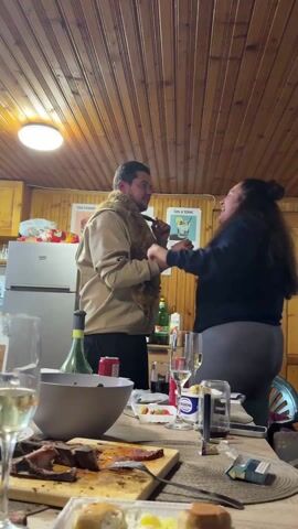 Two people are inside a room that appears to be a cabin. The walls and ceiling are made of wood planks. The room is filled with food, drinks, and dishes on a table. A man is wearing a tan sweatshirt and a fox pelt around his neck. The fox’s head is visible. A woman playfully grabs his hands and they laugh.