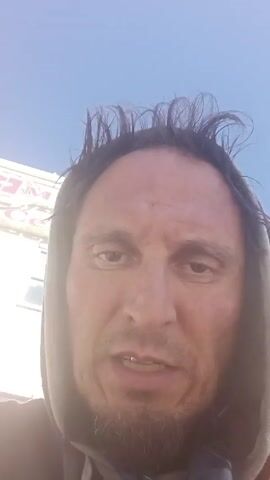 A man in a grey hoodie is filming a selfie-style video. His hair is somewhat messy and sticking up in places. He has a goatee and his expression appears serious as he speaks directly into the camera. His front teeth are visible. In the background, slightly out of focus, there's a white structure with some text that appears to say "Ice Cream". The sky is visible and is a clear blue.