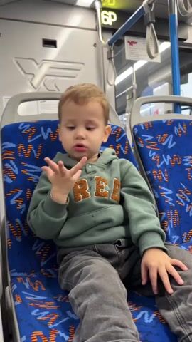 A young boy with blonde hair, wearing a green hoodie with the word "FREE" on it and grey pants, sits on a public transport seat with a blue and red patterned upholstery. The boy is gesturing with his hands, with his fingers moving in a playful manner. The background shows the interior of a bus or train, with seats, windows, and overhead luggage racks. A sign with "6P" is visible above the boy. The overall scene depicts a child interacting in a public transportation setting.