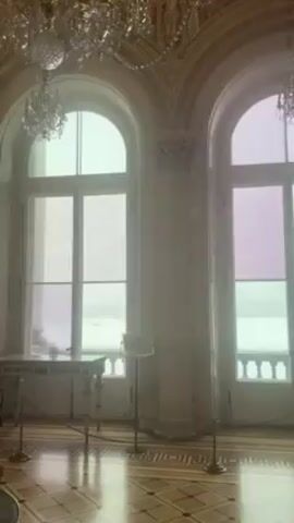The video shows the interior of an ornate room, likely within a museum or palace, featuring high, arched windows that offer a view of a bright, possibly overcast, outdoor scene. Two people wearing face masks walk into view, then past a large glass display case containing a gilded sculpture of a tree with birds. The room has a patterned wooden floor, decorative moldings on the walls and ceiling, and crystal chandeliers hanging from the ceiling. The display case has a gold frame and a red base, with guard rails.