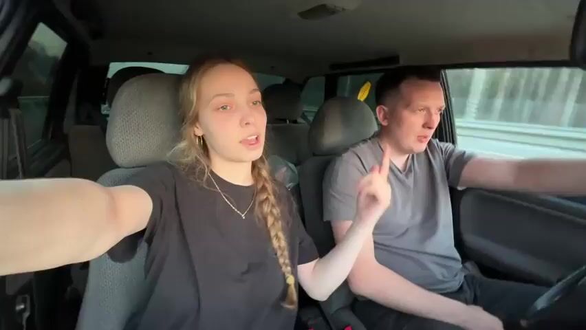 A woman with a braid and a man are sitting in a car. The man is driving. The woman is holding the camera and filming herself and the man. They are both wearing t-shirts. The car is driving on a highway. The woman is looking at the camera and talking. The man is looking at the road. There is music playing in the background.
