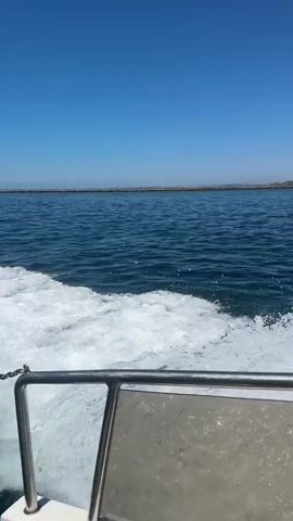 The video is filmed from the perspective of someone on a boat moving through the water. The camera is positioned low, looking forward and slightly to the side. A metal railing is visible in the foreground, partially obstructing the view of the water. White foam and spray from the boat's wake are prominent on the left side of the frame. In the distance, a coastline with buildings and a beach can be seen under a clear blue sky. Another smaller boat with people on board is visible on the water further away, moving in the same general direction as the camera's boat. The water is a deep blue.