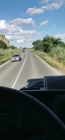 The video shows a first-person perspective from inside a vehicle, likely a truck, as it drives down a paved road. The road is in good condition with a white lane divider. On the left side of the road, there is a steep, grassy embankment with some rocks and vegetation. On the right side, there are fields of tall green grass and trees. The sky is blue with scattered white clouds. In the distance, there are a few other vehicles, including a car and possibly a truck. The sun appears to be shining brightly, suggesting it is daytime. The overall scene depicts a rural or suburban landscape during fair weather.