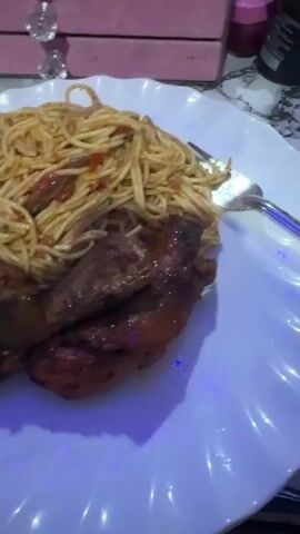 A plate of food with spaghetti and fried plantains is centered in the frame. The spaghetti appears to have a tomato-based sauce. Three pieces of plantain sit beneath the spaghetti. A fork is present on the right side of the plate. The plate itself is white with a scalloped edge. The lighting on the table appears to be blue, causing shadows on the plate. A pink storage container with diamond accents is visible in the background, along with a partial view of a decorative glass. The camera angle shifts slightly during the video.