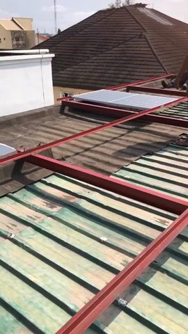This video captures a rooftop scene in Ilado, Nigeria, likely during daytime. The focus is on the installation of solar panels on a corrugated metal roof. Two men are present, actively engaged in the task. One man, wearing a pink polo shirt and grey jeans, stands on the roof, seemingly directing or supervising. Another man, in a lighter colored shirt and dark pants, is seen further along the roof, also participating in the installation.

The scene is dominated by the metallic sheen of the roof and the bright red metal framework being erected to support the solar panels. Several solar panels are already in place, glinting in the sunlight. In the background, residential buildings with tiled roofs are visible, along with water tanks mounted on a structure. The sky appears bright, suggesting a clear or partly cloudy day.

The activity clearly depicted is the assembly and mounting of solar panels, a process indicative of a renewable energy project. The presence of the men working on the roof, the materials laid out, and the partially installed panels all point towards an ongoing construction or installation event. The overall atmosphere seems industrious, with the workers focused on their task under the Nigerian sun. There is no visible text in the video.
