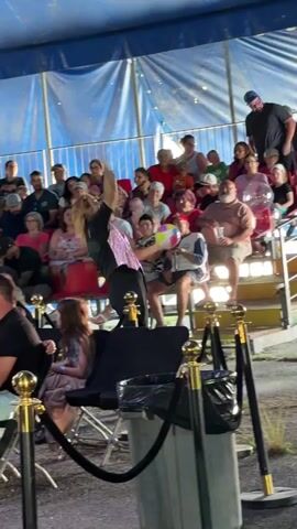 A person with blonde hair wearing a black shirt and black pants is performing in front of an audience seated on bleachers. The performer is holding a colorful ball in their left hand and appears to be interacting with the audience. The audience members are diverse in age and gender. The environment appears to be under a tent with blue and white striped fabric. There are gold stanchions with red ropes in the foreground, suggesting a restricted area. A trash can is also visible in the foreground. The lighting suggests an outdoor daytime event.