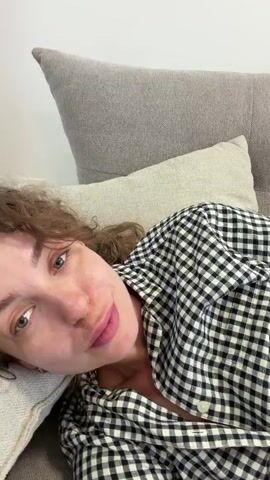 A person with curly brown hair, wearing a black and white checkered shirt, lies on a gray sofa with a pillow behind their head. The person is looking at the camera with a slight smile. A light brown, fluffy dog is positioned next to the person on the sofa. The dog is lying on its back with its legs in the air and its face partially visible. The video is filmed from a close-up perspective.
