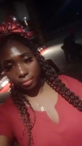 A young woman with dark skin and a red outfit is taking a selfie outside at night. She has long, dark braids and is wearing a red bow headband. She is also wearing a gold necklace. There are other people in the background, but they are out of focus. Loud music is playing.