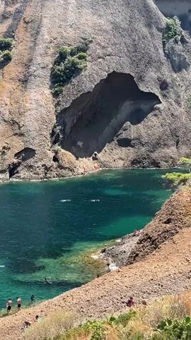 A video taken from an elevated viewpoint shows a natural inlet or cove with steep, rocky cliffs on either side and a cave opening in the far cliff face. The water is clear and turquoise, with visible rocks beneath the surface near the shore. Several people are seen in the water, swimming and enjoying the day. On the rocky shoreline to the right, a group of people are gathered. Further down the shoreline, near the water's edge, are two kayaks, one red and yellow, the other yellow and red, resting on the rocks. The sun is shining, casting shadows and highlighting the textures of the rock formations.