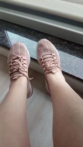 The video shows a person's legs and feet wearing light pink athletic shoes. The shoes are laced up and have a mesh-like texture. The person is sitting or resting, with their legs extended forward. The floor appears to be light-colored wood or laminate. In the background, there is a dark, speckled surface, possibly a countertop or windowsill, and a window with horizontal blinds. Sunlight streams through the blinds, casting shadows on the surface. The scene suggests an indoor setting, possibly a home or an office, with natural light. The activity is static, with no visible movement beyond slight shifts in leg position. The overall impression is casual and relaxed. There is no visible text. Given the context of Ankara, Turkey, the time of day could be daytime, and the weather is likely sunny or bright, indicated by the strong light.